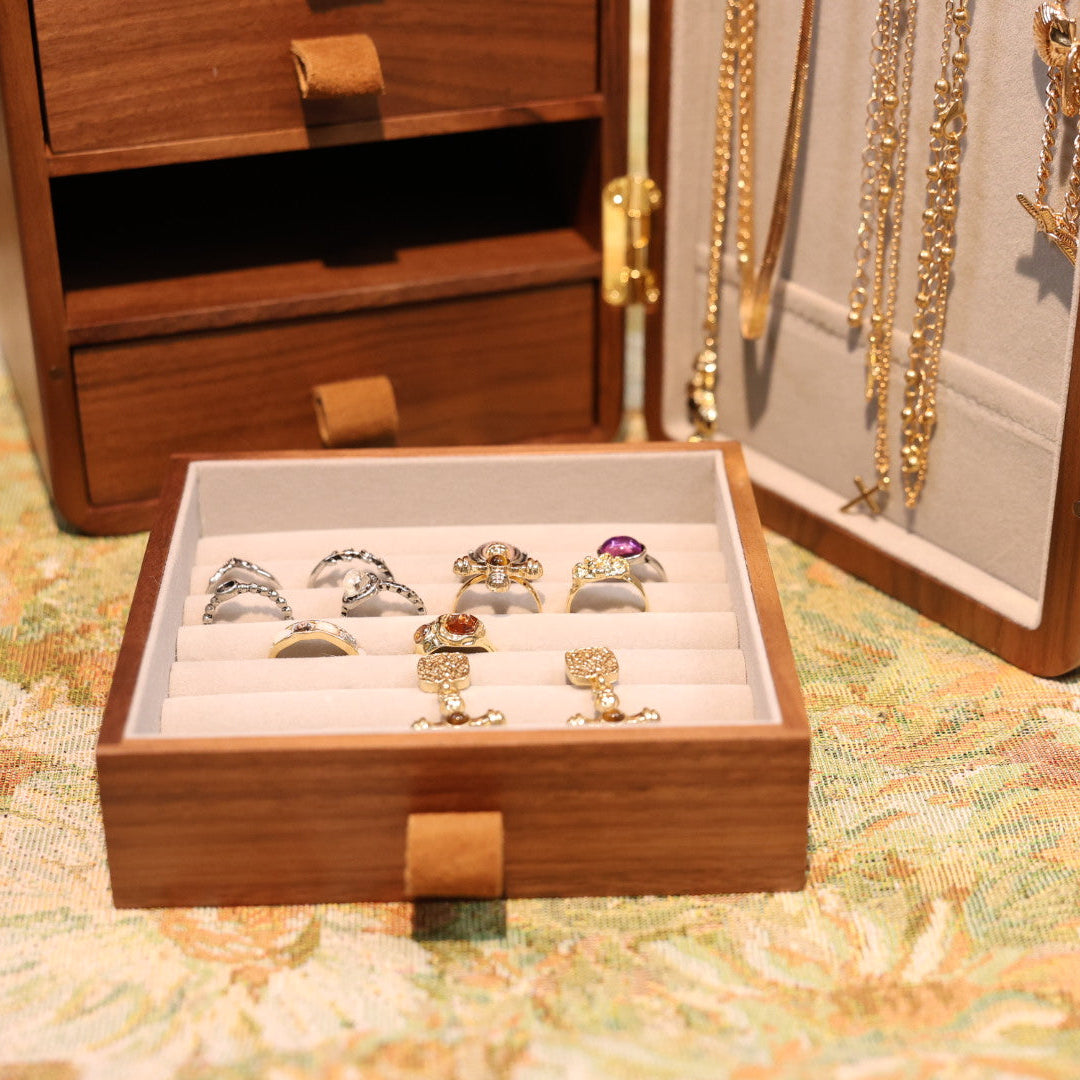 Wooden jewelry box with open lid displaying necklaces and a tray with rings on a floral tablecloth.