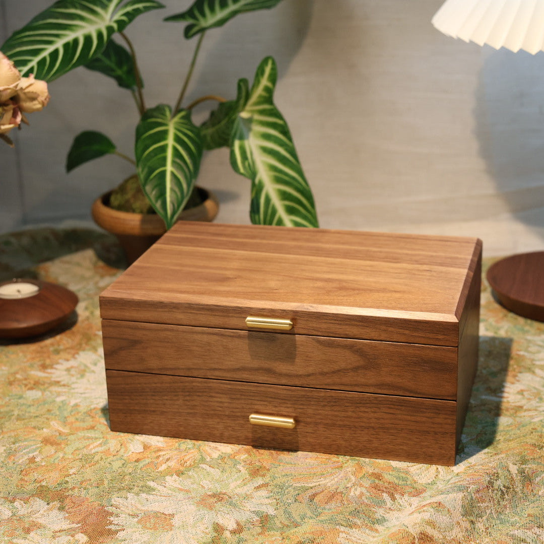 Wooden jewelry box on a patterned surface with decorative plants and a lamp in the background.
