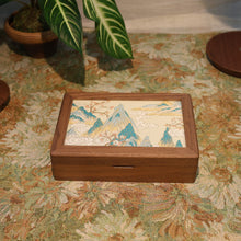 Wooden box with a decorative lid featuring mountains on a floral-patterned tablecloth.
