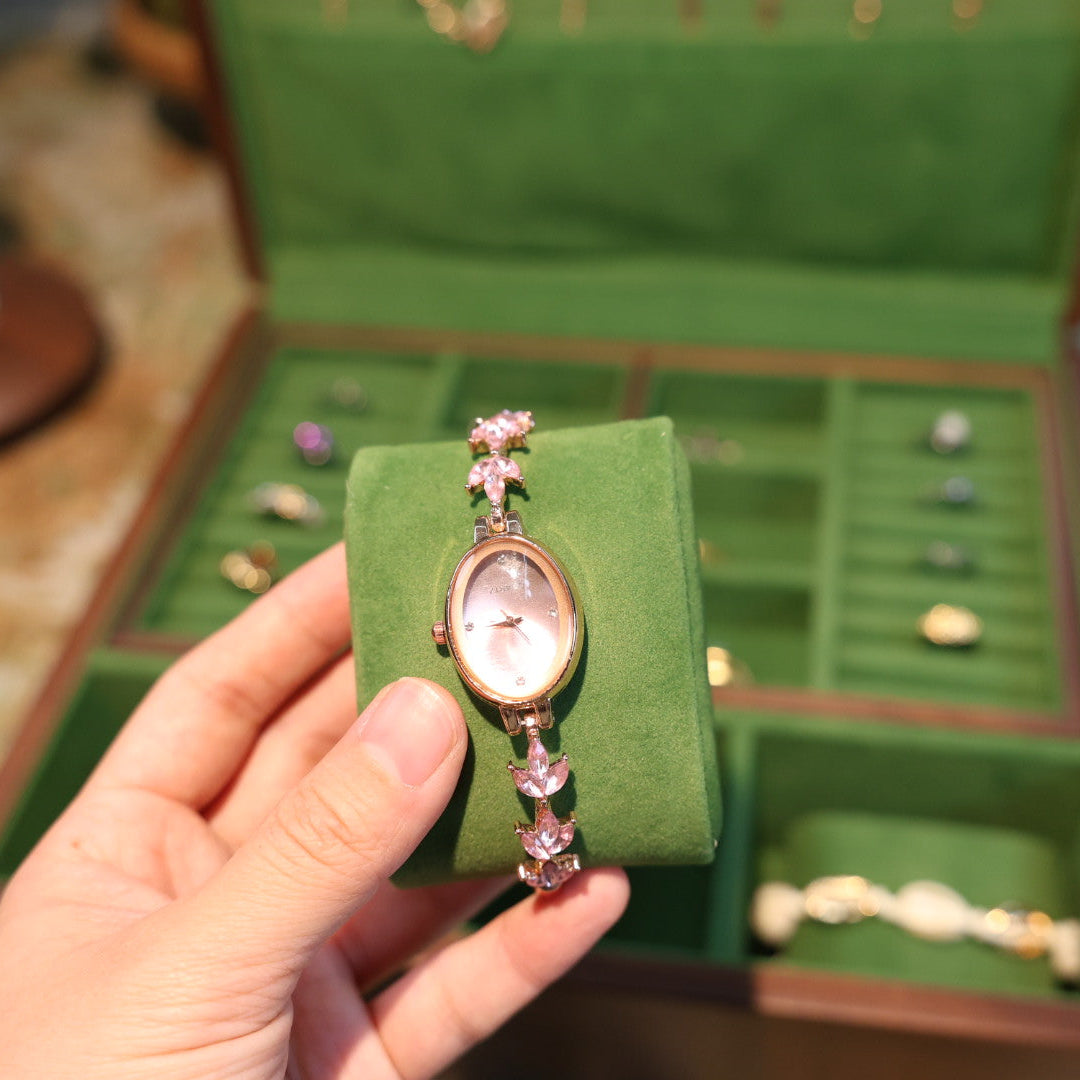 Hand holding a watch with pink gemstones in front of an open green jewelry box.