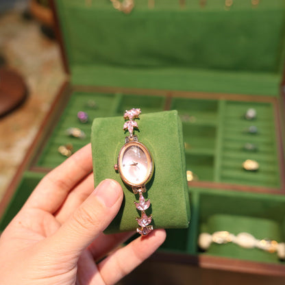 Hand holding a watch with pink gemstones in front of an open green jewelry box.